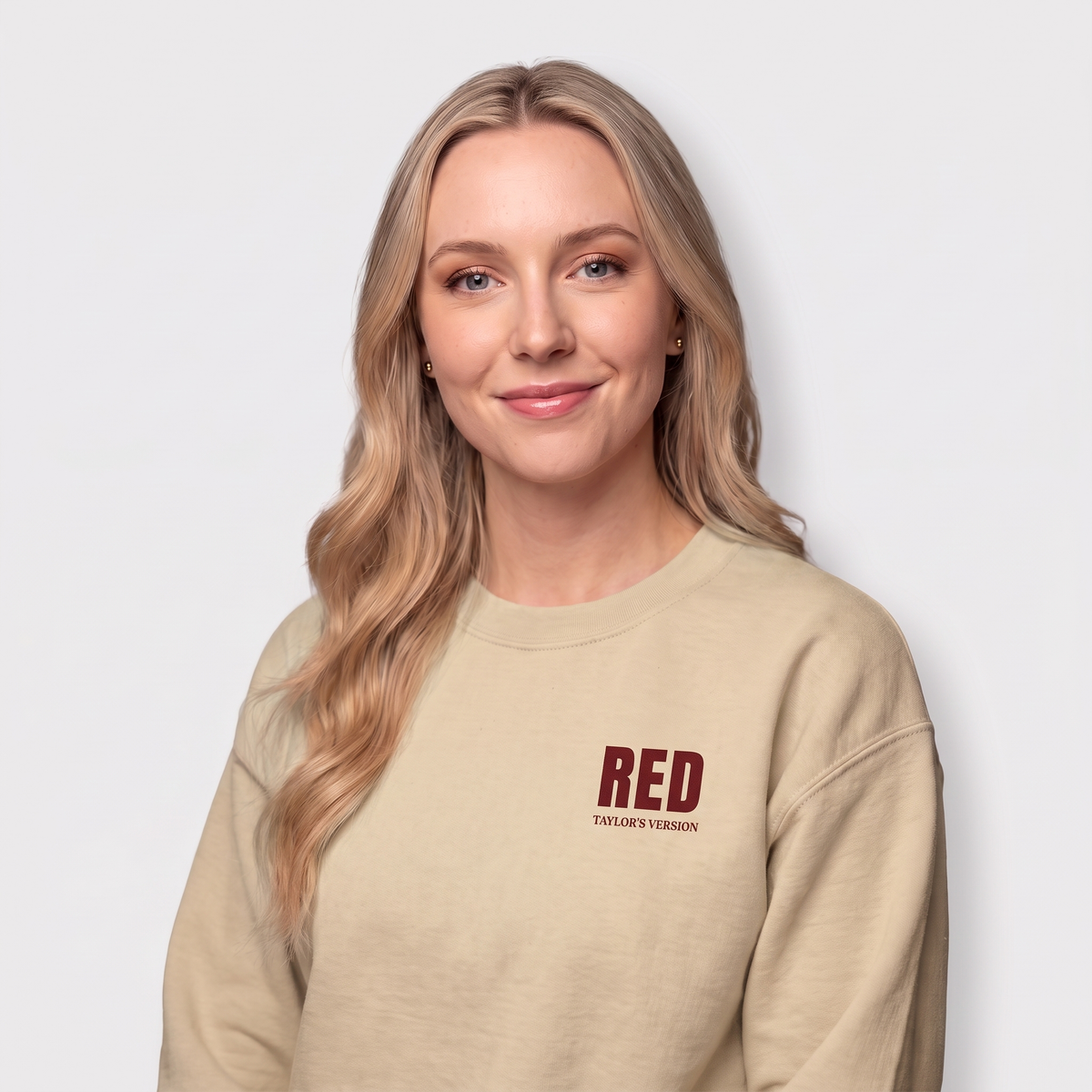 A woman with blonde hair wearing a beige crewneck sweatshirt with a dark red graphic on the left chest that reads 'RED TAYLOR'S VERSION', set against a plain light gray background.