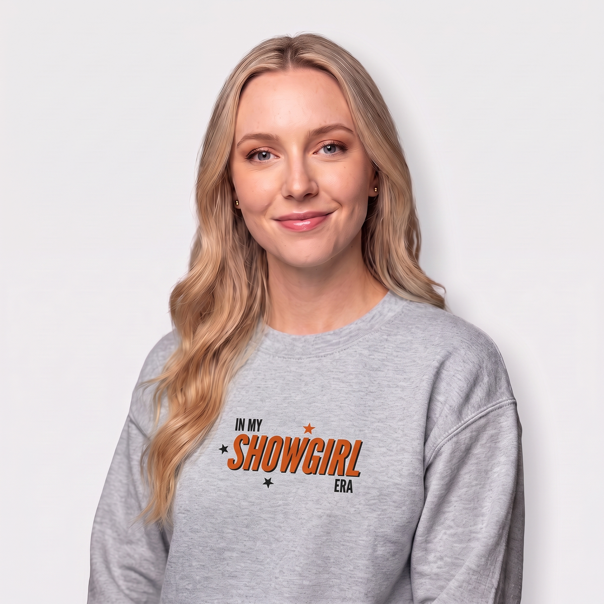 A blonde woman wearing a heather gray crewneck sweatshirt with a graphic that reads 'IN MY SHOWGIRL ERA' in orange and black, photographed against a light gray studio background.