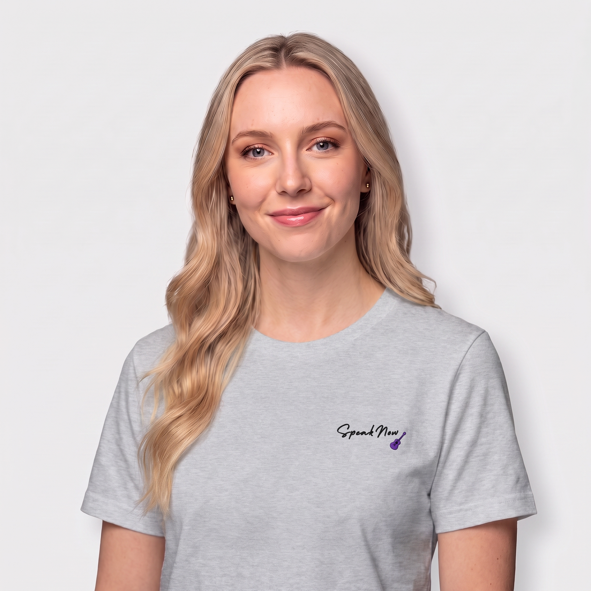 A blonde woman wearing a light gray crewneck t-shirt with a small black 'Speak Now' script and purple flower graphic on the left chest, set against a plain light gray background.