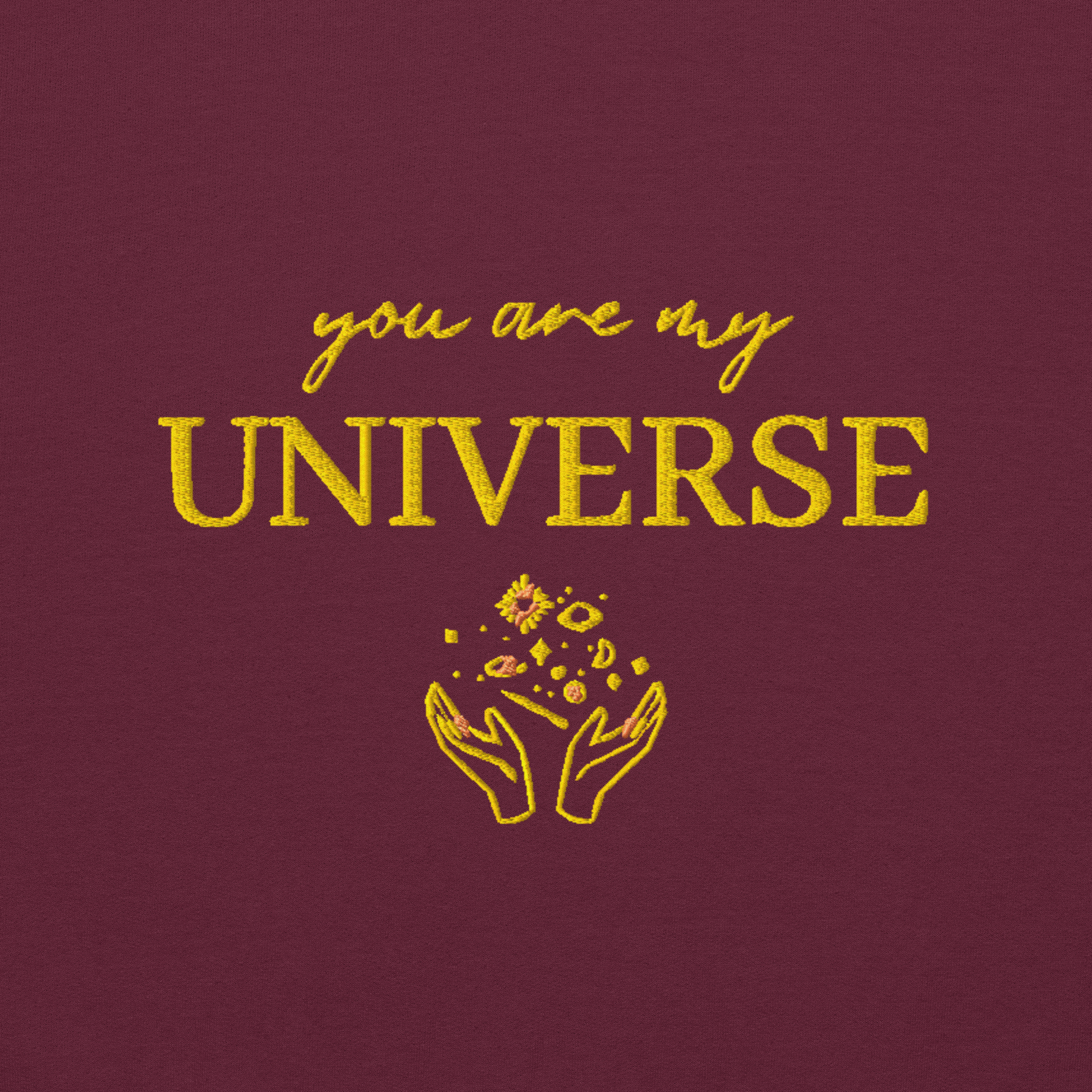 Maroon crewneck sweatshirt featuring yellow typography that reads “you are my Universe” with minimalist hands holding stars illustration -inspired by Coldplay’s hit song “My Universe”, aesthetic fan apparel for music lovers.