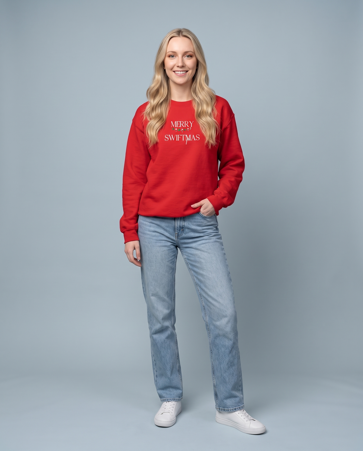A woman modeling a red crewneck sweatshirt with 'MERRY CHRISTMAS' printed in white on the chest, paired with light blue jeans and white sneakers, against a solid gray studio background.