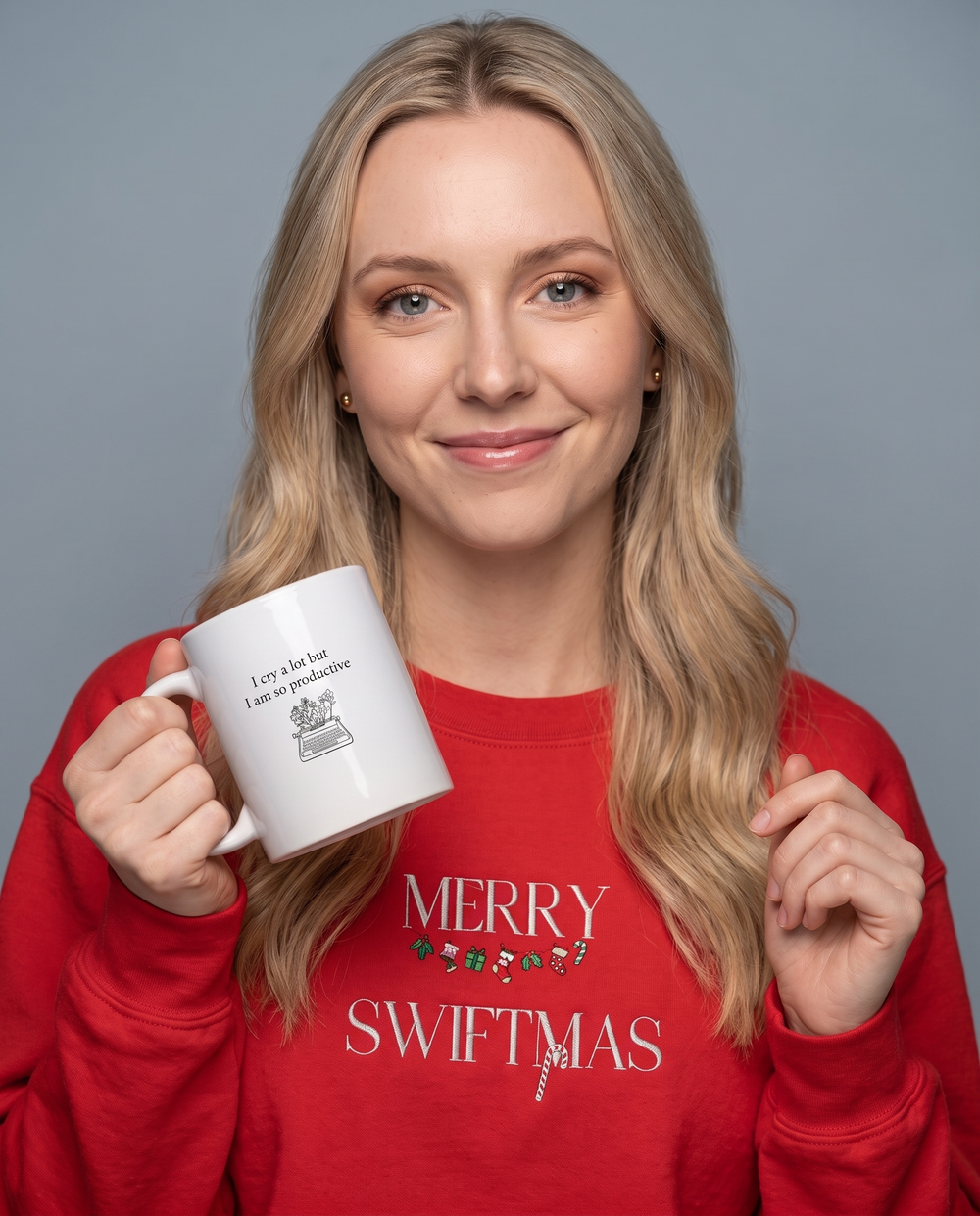 A woman with blonde hair wearing a red sweatshirt with 'MERRY SWIFTMAS' printed on it, holding a white ceramic mug that says 'I cry a lot but I smile whenever'. She is posed against a solid gray studio background.