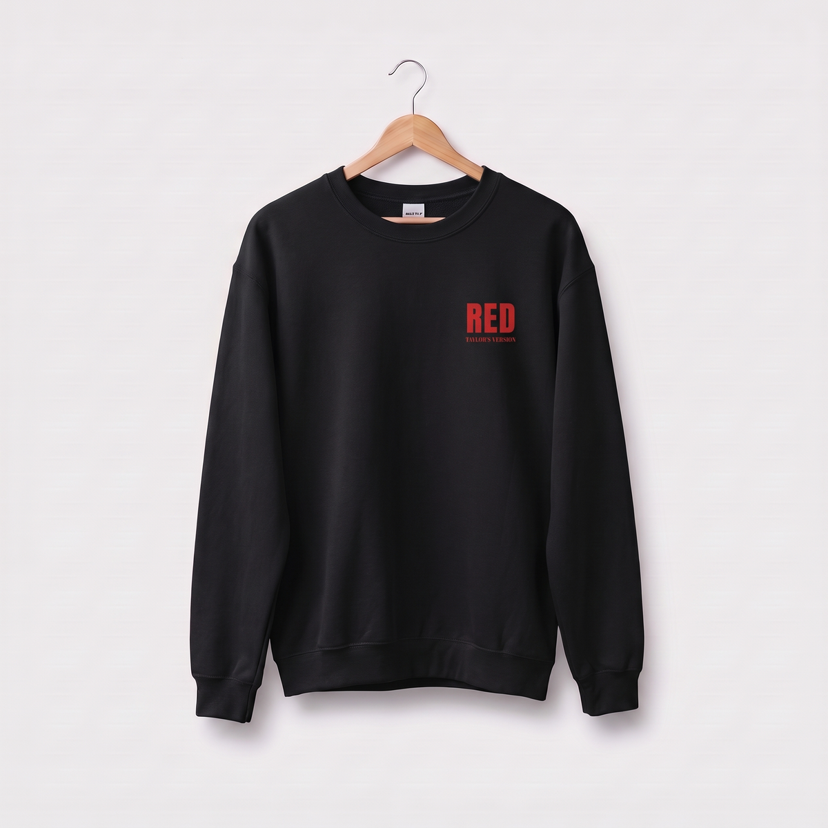 A navy blue crewneck sweatshirt hanging on a wooden hanger against a light gray background. The sweatshirt features a small red graphic on the left chest that reads 'RED' with 'TAYLOR'S VERSION' underneath.