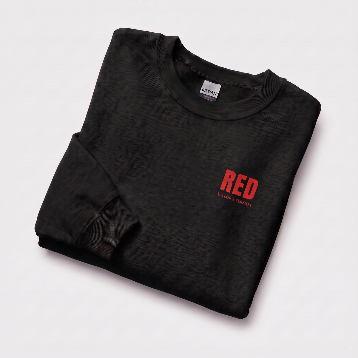 A folded black crewneck sweatshirt with a subtle textured pattern, featuring a red graphic print on the left chest that reads 'RED' with 'TAYLOR'S VERSION' underneath, displayed on a clean white background.