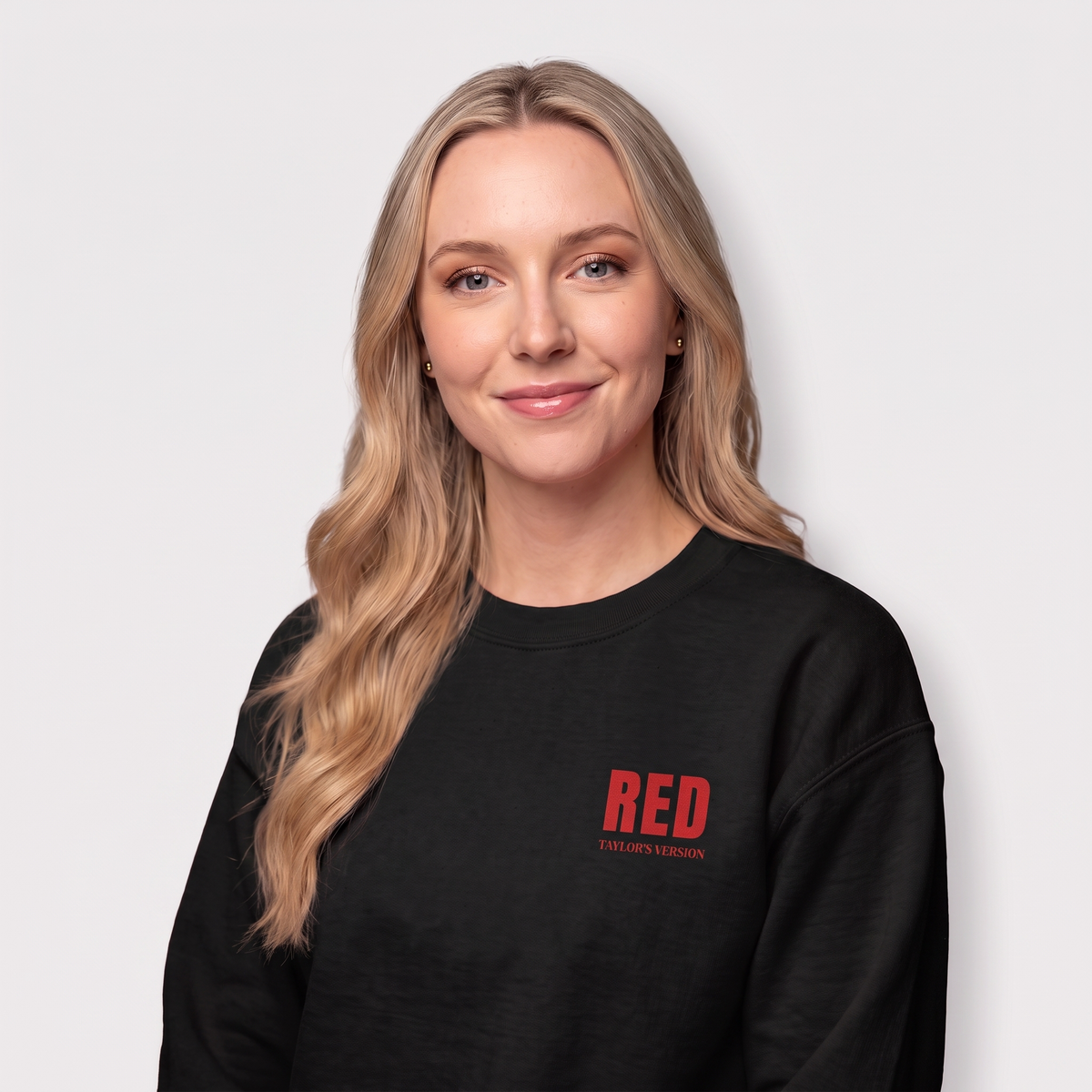 A woman with blonde hair wearing a black crewneck sweatshirt with 'RED TAYLOR'S VERSION' printed in red on the left chest, set against a plain light gray background.