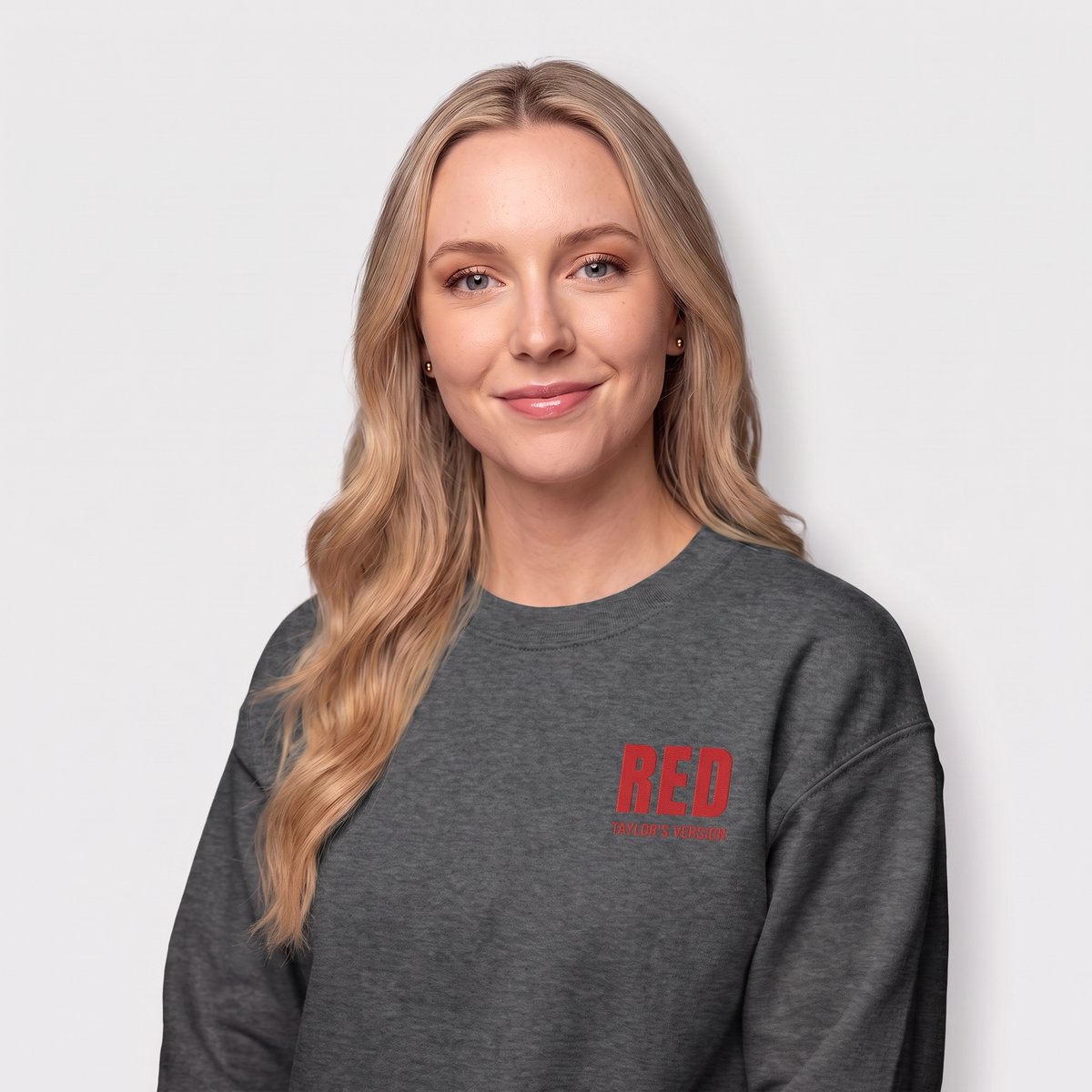 A woman with blonde hair wearing a dark gray crewneck sweatshirt with a red 'RED TAYLOR'S VERSION' logo on the left chest, photographed against a light gray background.