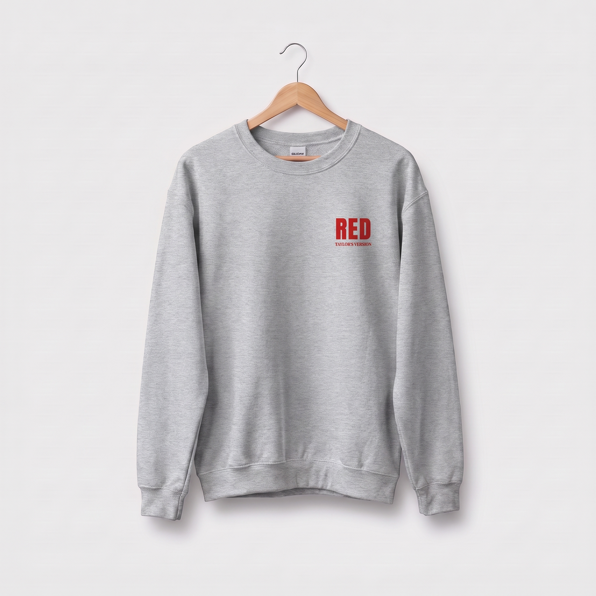 A light gray crewneck sweatshirt hanging on a wooden hanger against a light gray background. The sweatshirt features a small red graphic on the left chest that reads 'RED' with 'TAYLOR'S VERSION' underneath.