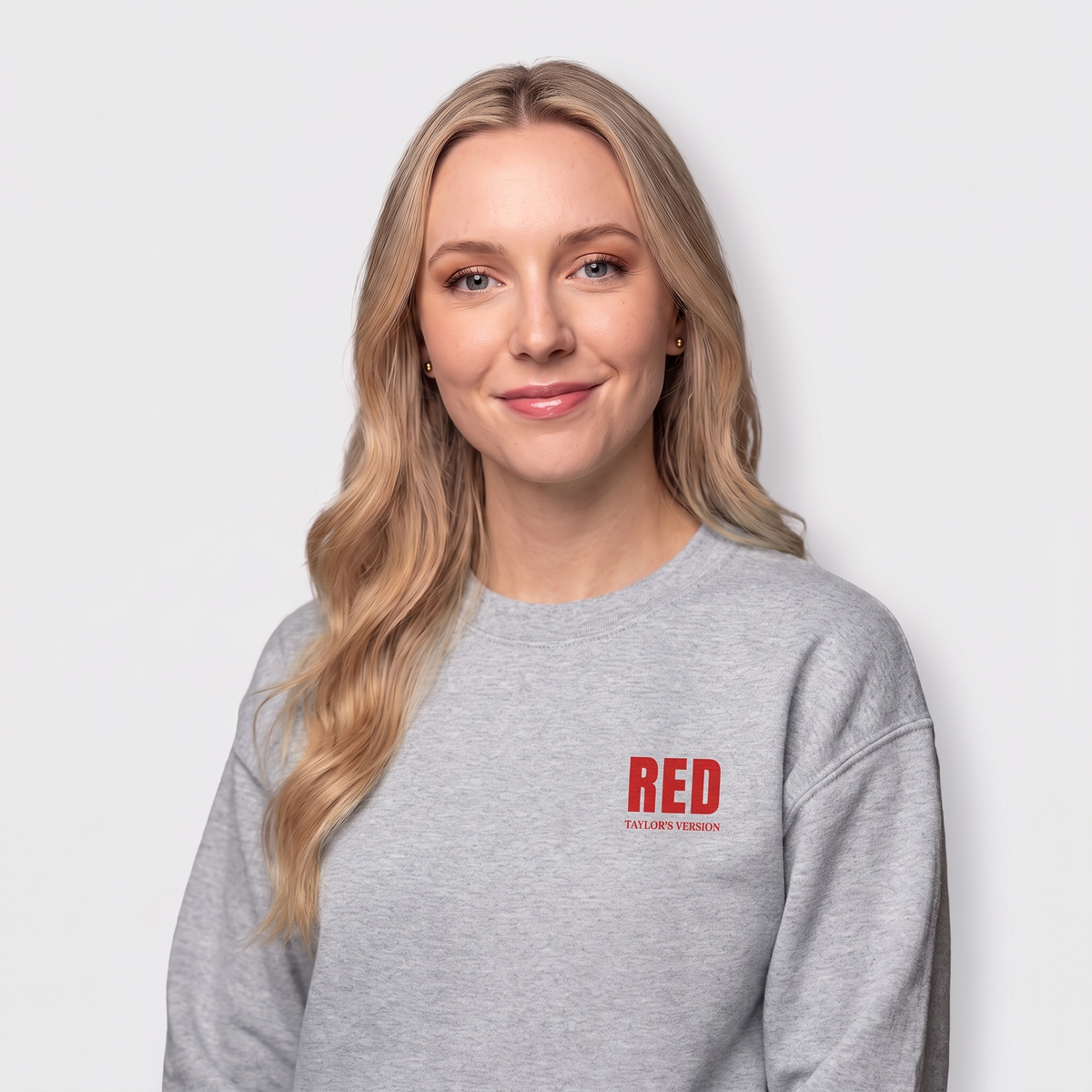 A blonde woman wearing a heather gray crewneck sweatshirt with a red 'RED' logo on the left chest, photographed against a light gray studio background.