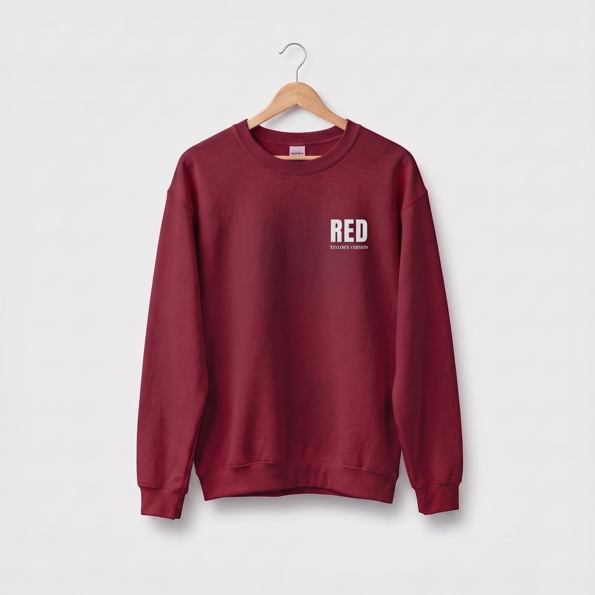 A maroon crewneck sweatshirt hanging on a wooden hanger against a light gray background. The sweatshirt features a small white graphic on the left chest that reads 'RED' with 'TAYLOR'S VERSION' underneath.