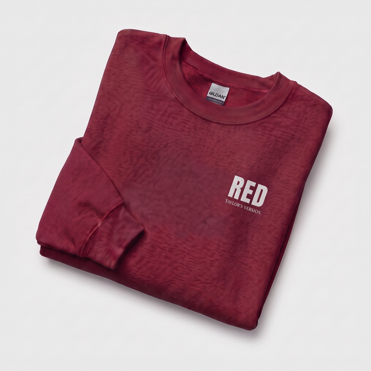 A folded long-sleeve crewneck sweatshirt in a heathered dark red color, featuring a white graphic print on the left chest that reads 'RED' with 'TAYLOR'S VERSION' underneath. The brand 'Gildan' is visible on the inner neck label.