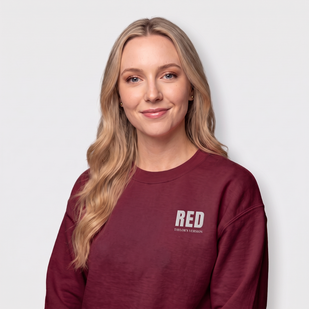 A woman with long blonde hair wearing a maroon crewneck sweatshirt with 'RED' and 'TAYLOR'S VERSION' printed in white on the left chest, set against a plain light gray background.