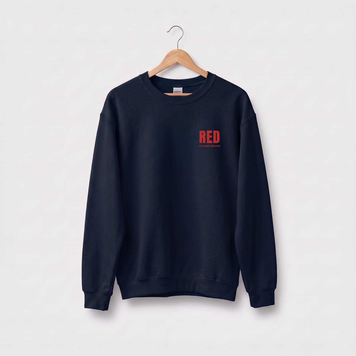 A navy blue crewneck sweatshirt hanging on a wooden hanger against a light gray background. The sweatshirt features a small red graphic on the left chest that reads 'RED' with 'TAYLOR'S VERSION' underneath.