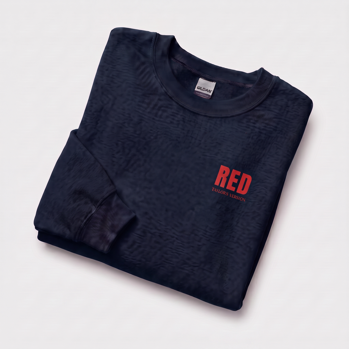 A folded dark navy crewneck sweatshirt with a subtle textured pattern, featuring a red 'RED' graphic and smaller 'TAYLOR'S VERSION' text on the left chest, set against a light off-white background.