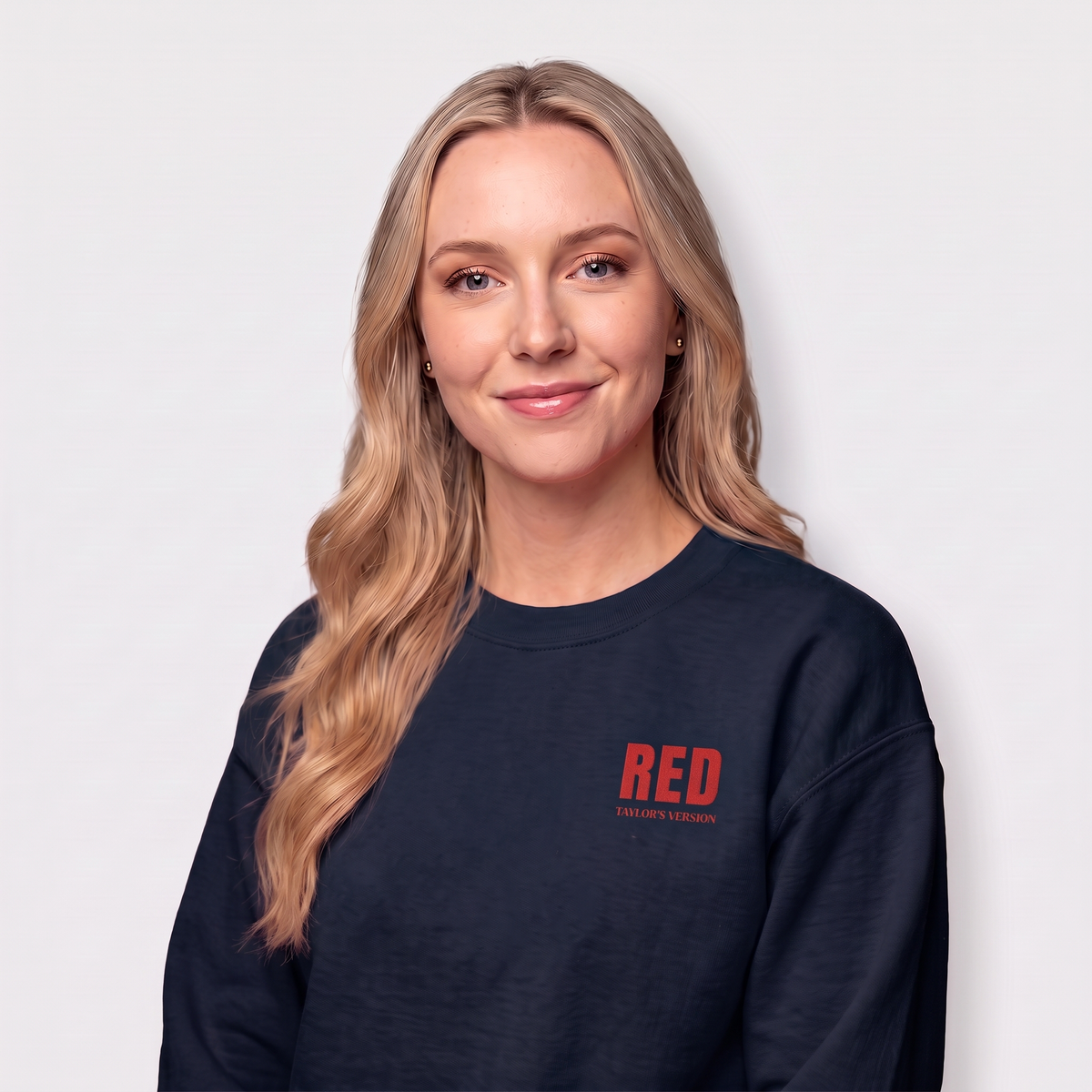 A woman with blonde hair wearing a dark navy crewneck sweatshirt with a red 'RED' TAYLOR'S VERSION' logo on the left chest, photographed against a light gray background.