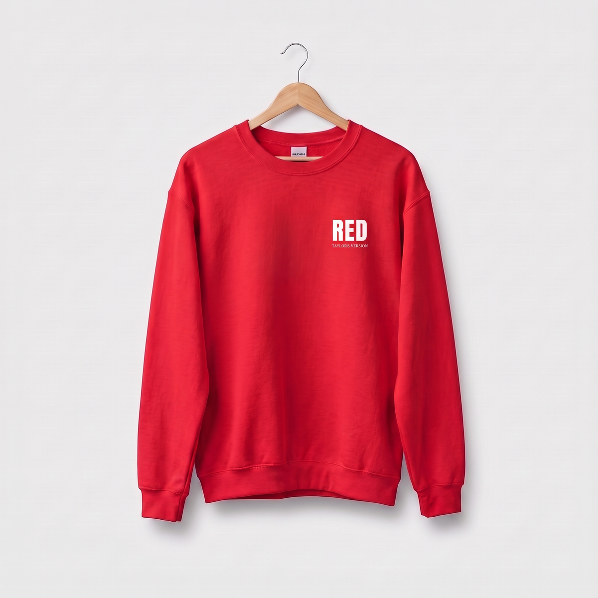 A bright red crewneck sweatshirt hanging on a wooden hanger against a light gray background. The sweatshirt features a small white graphic on the left chest that reads 'RED' with 'TAYLOR'S VERSION' printed underneath.