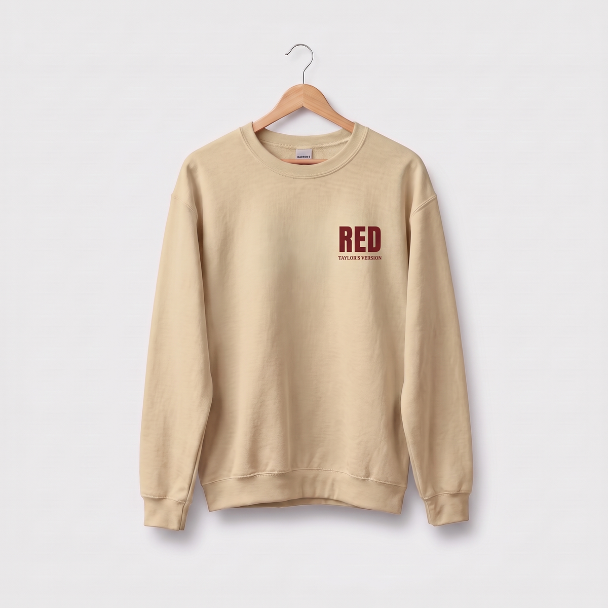 A beige crewneck sweatshirt hanging on a wooden hanger against a light gray background, featuring a dark red 'RED' graphic on the left chest with 'Taylor's Version' printed underneath.