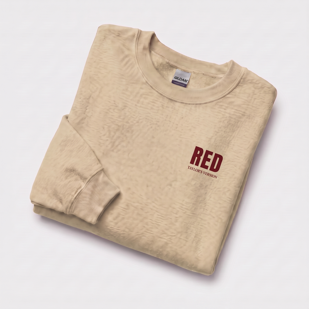 A folded beige crewneck sweatshirt with a small red graphic on the left chest reading 'RED TAYLOR'S VERSION', displayed on a clean white background.