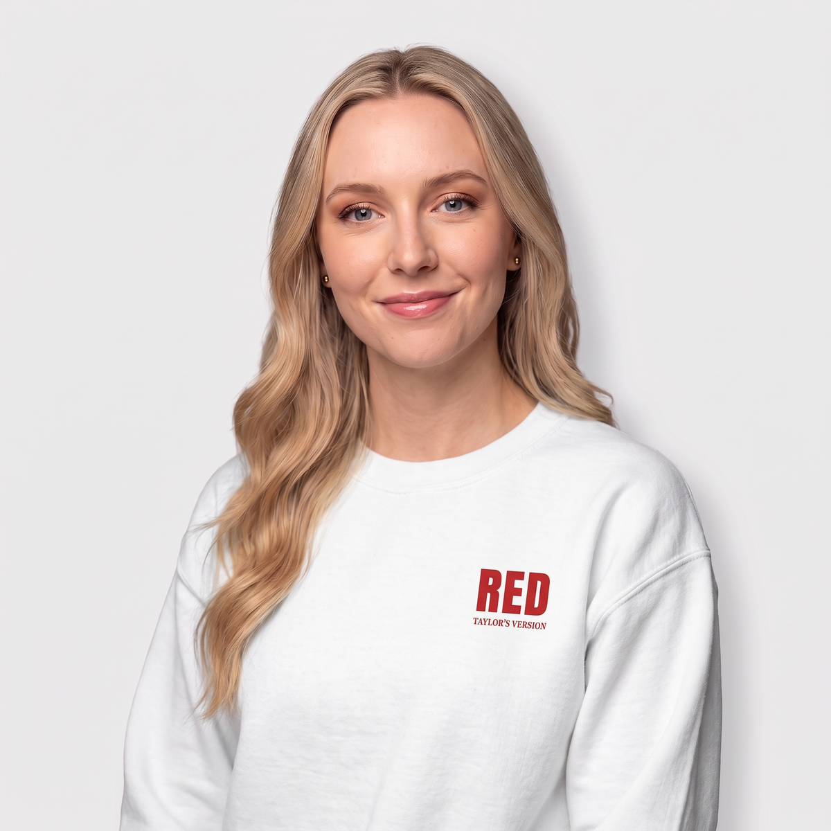 A blonde woman wearing a white crewneck sweatshirt with a red 'RED' logo and 'TAYLOR'S VERSION' text on the left chest, photographed against a light gray background.