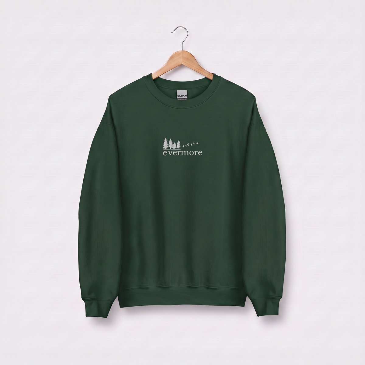 Forest green Evermore sweatshirt inspired by Taylor Swift's evermore album, embroidered pine tree and flying birds logo, cottagecore Swiftie merch on a wooden hanger