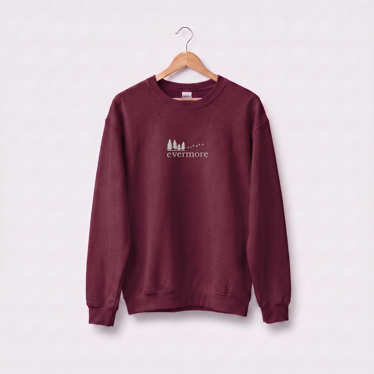 Maroon Evermore sweatshirt inspired by Taylor Swift's evermore album, embroidered pine tree and flying birds logo, cottagecore Swiftie merch on a wooden hanger