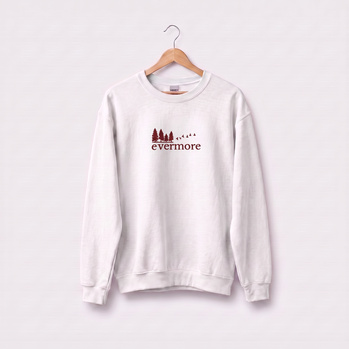 White Evermore sweatshirt inspired by Taylor Swift's evermore album, embroidered pine tree and flying birds logo, cottagecore Swiftie merch on a wooden hanger