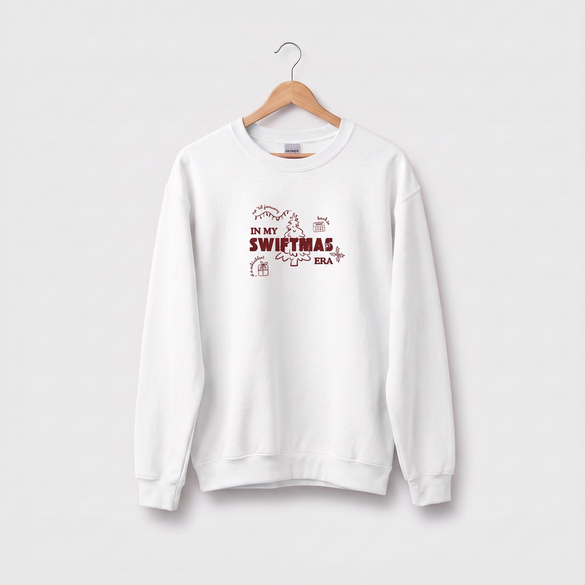 A white crewneck sweatshirt hanging on a wooden hanger against a light gray background. The sweatshirt features a graphic print on the chest with the text 'IN MY SWITMAS ERA' in dark red, accompanied by small illustrations of Christmas trees and ornaments.