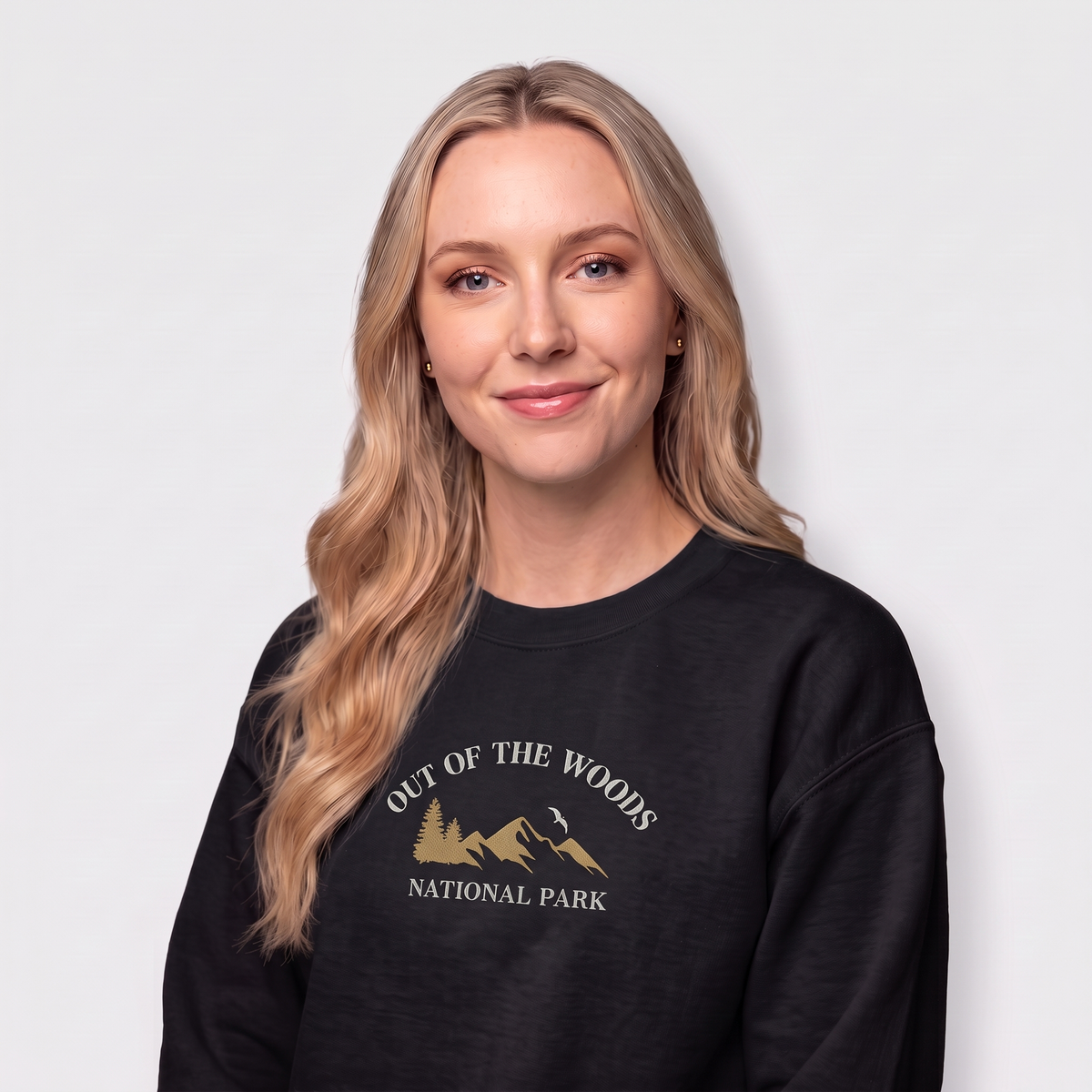 A woman with long blonde hair wearing a black crewneck sweatshirt with a gold graphic of mountains and trees and the text 'OUT OF THE WOODS NATIONAL PARK' on the front, set against a plain light gray background.