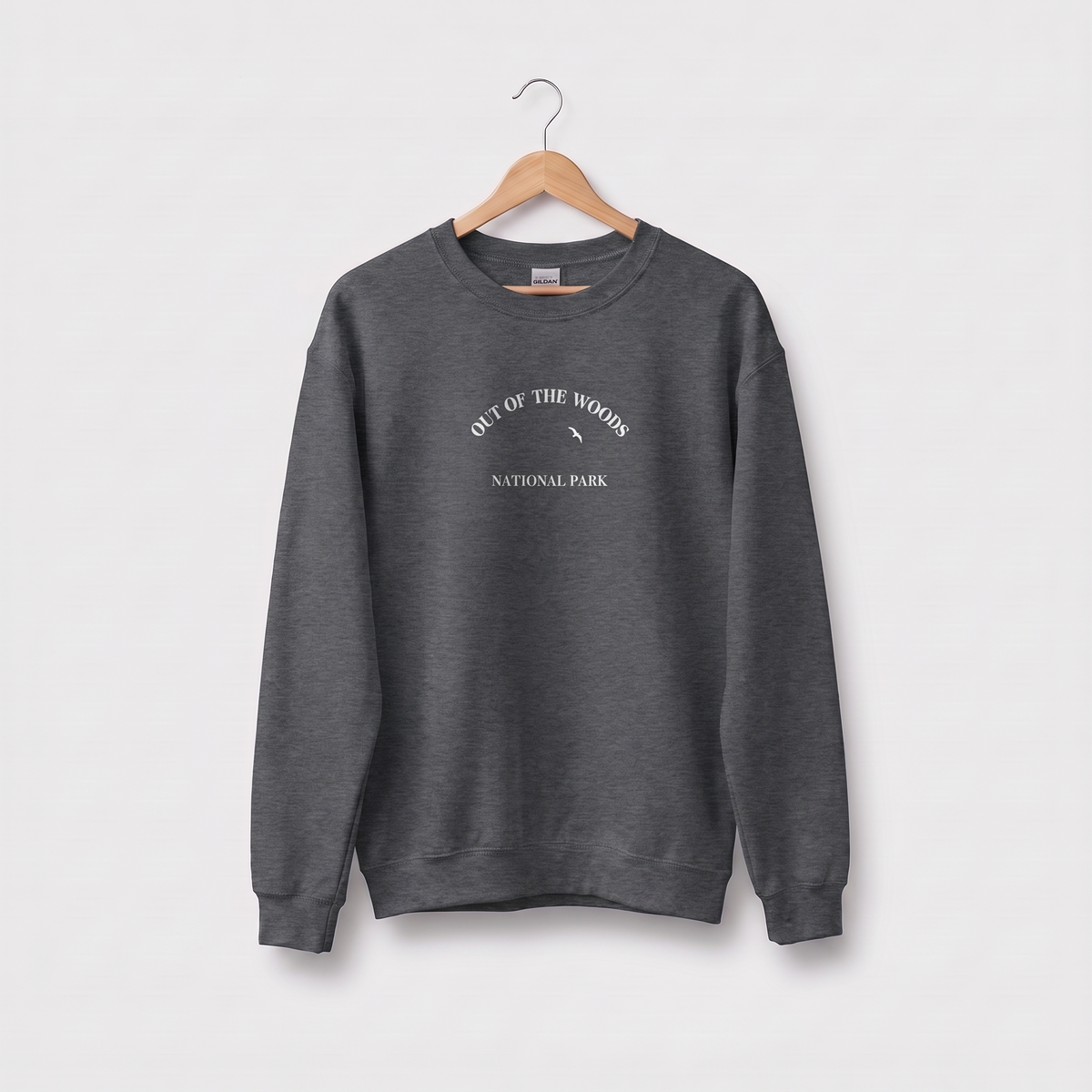 A dark heather crewneck sweatshirt hanging on a wooden hanger against a light gray background. The sweatshirt features a graphic on the chest reading 'OUT OF THE WOODS NATIONAL PARK' in white text with a mountain illustration.