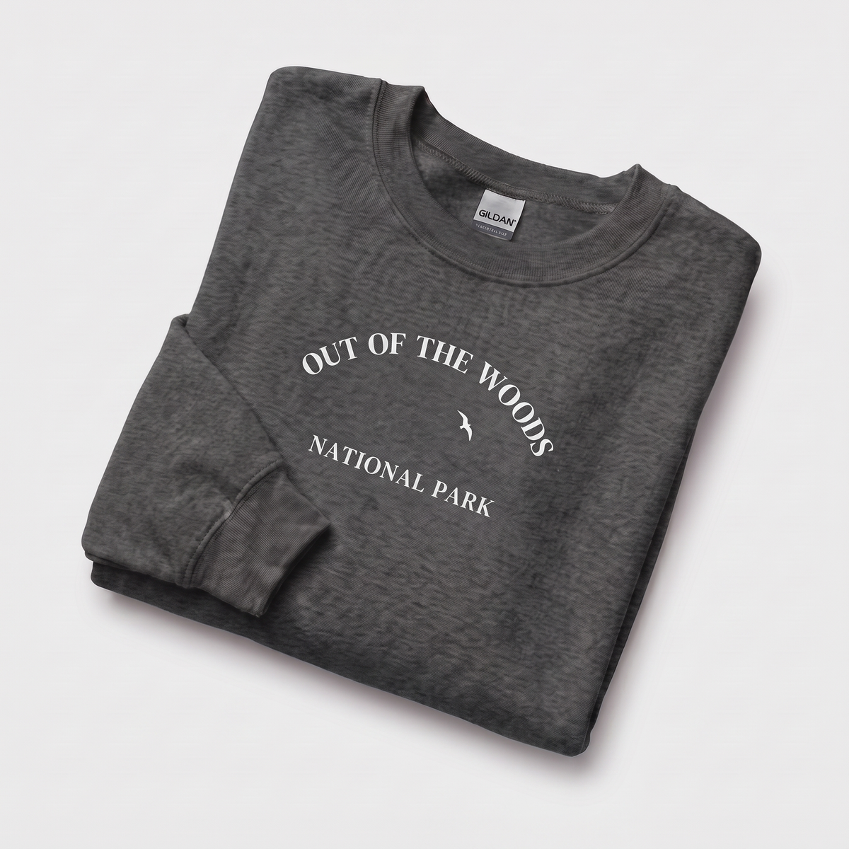 A folded crewneck sweatshirt in a heathered dark navy, featuring a white  graphic print of a mountain and trees with the text 'OUT OF THE WOODS NATIONAL PARK'. A Gildan brand tag is visible inside the collar.