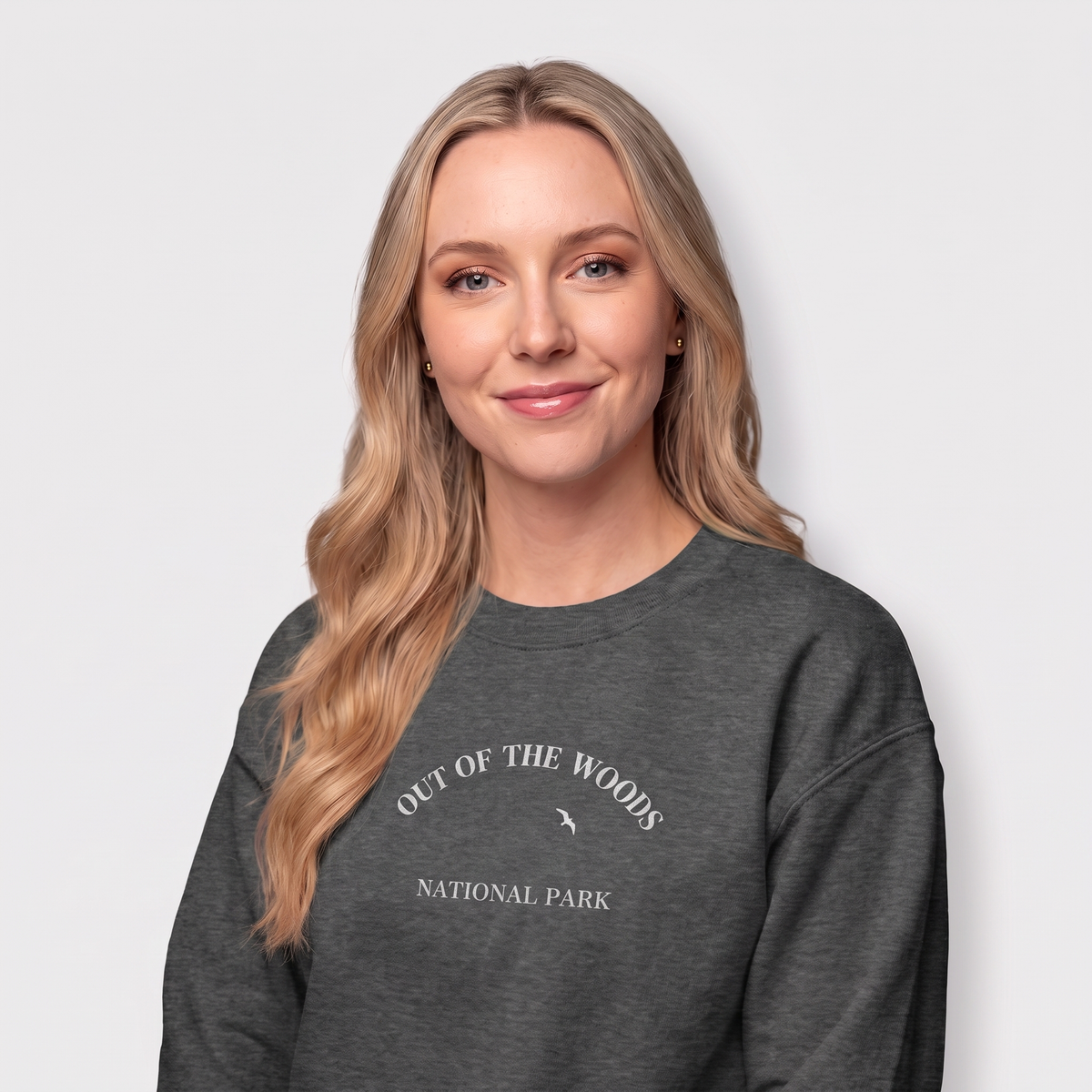 A woman with long blonde hair wearing a dark heather crewneck sweatshirt with a white graphic of mountains, trees, and the text 'OUT OF THE WOODS NATIONAL PARK' on the front, set against a light gray background.