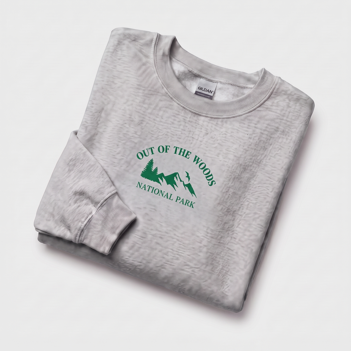 A folded light gray heather crewneck sweatshirt with a green graphic print reading 'OUT OF THE WOODS NATIONAL PARK' and a tree-and-person silhouette, shown on a clean white background. A Gildan neck label is visible.