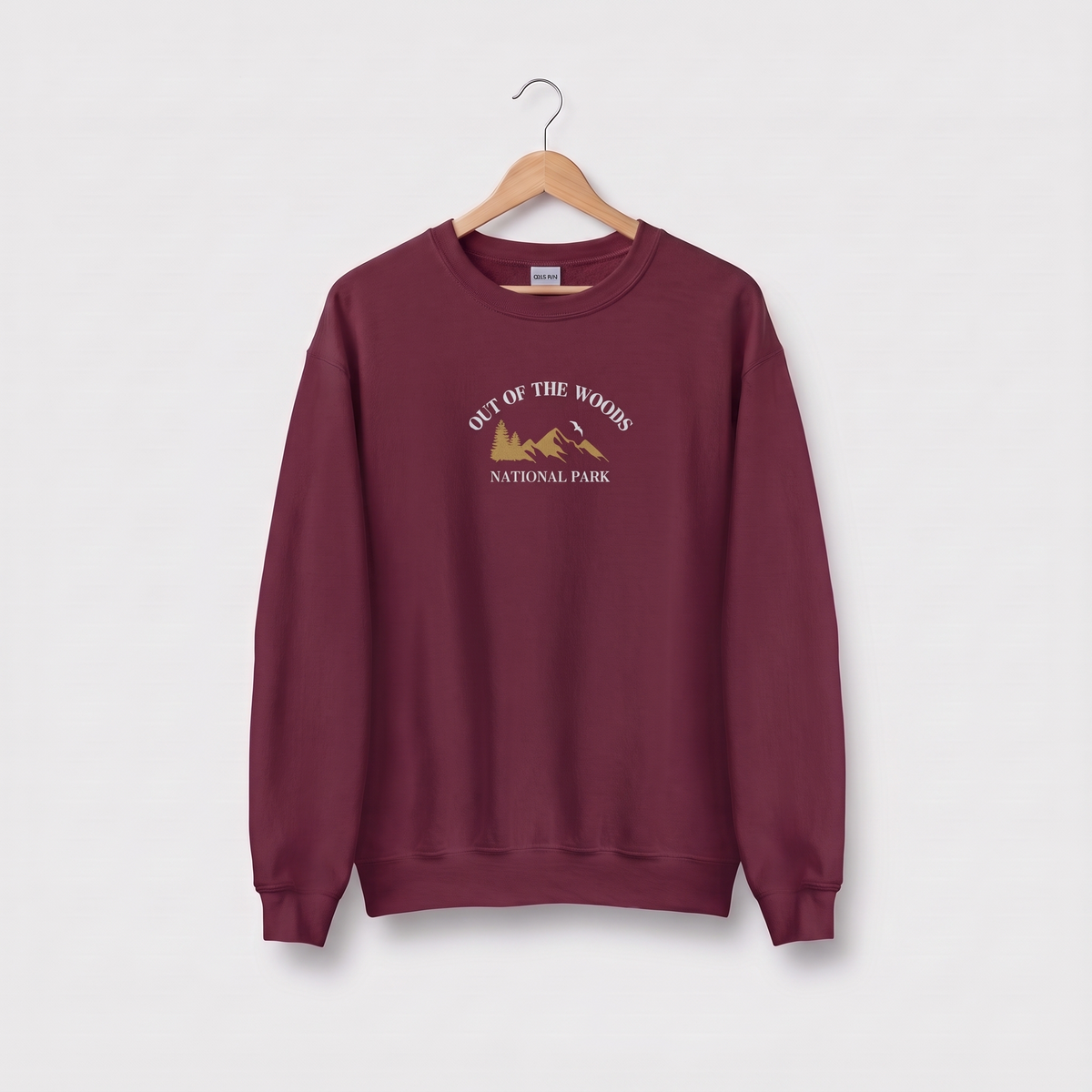 A maroon crewneck sweatshirt hanging on a wooden hanger against a light gray background. The sweatshirt features a graphic on the chest reading 'OUT OF THE WOODS NATIONAL PARK' in white text with a gold mountain illustration.