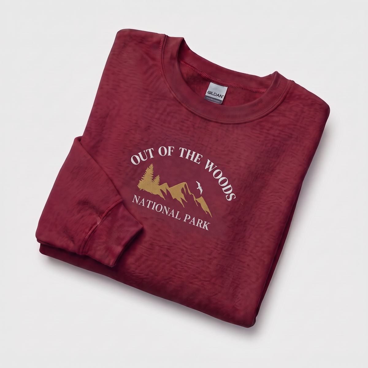 A folded crewneck sweatshirt in a heathered burgundy red, featuring a gold and white graphic print of a mountain and trees with the text 'OUT OF THE WOODS NATIONAL PARK'. A Gildan brand tag is visible inside the collar.