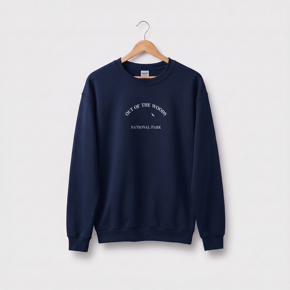 A navy blue crewneck sweatshirt hanging on a wooden hanger against a light gray background. The sweatshirt features a white graphic print on the chest that reads 'OUT OF THE WOODS NATIONAL PARK' with a small tree silhouette.