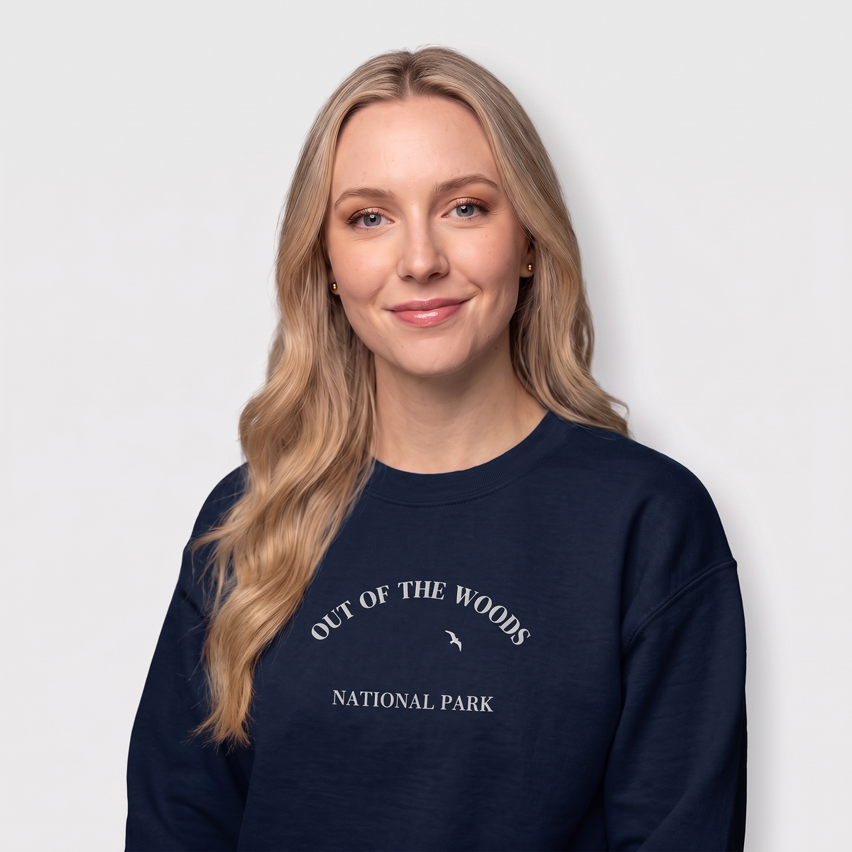 A woman with long blonde hair wearing a navy crewneck sweatshirt with a white graphic of mountains, trees, and the text 'OUT OF THE WOODS NATIONAL PARK' on the front, set against a light gray background.