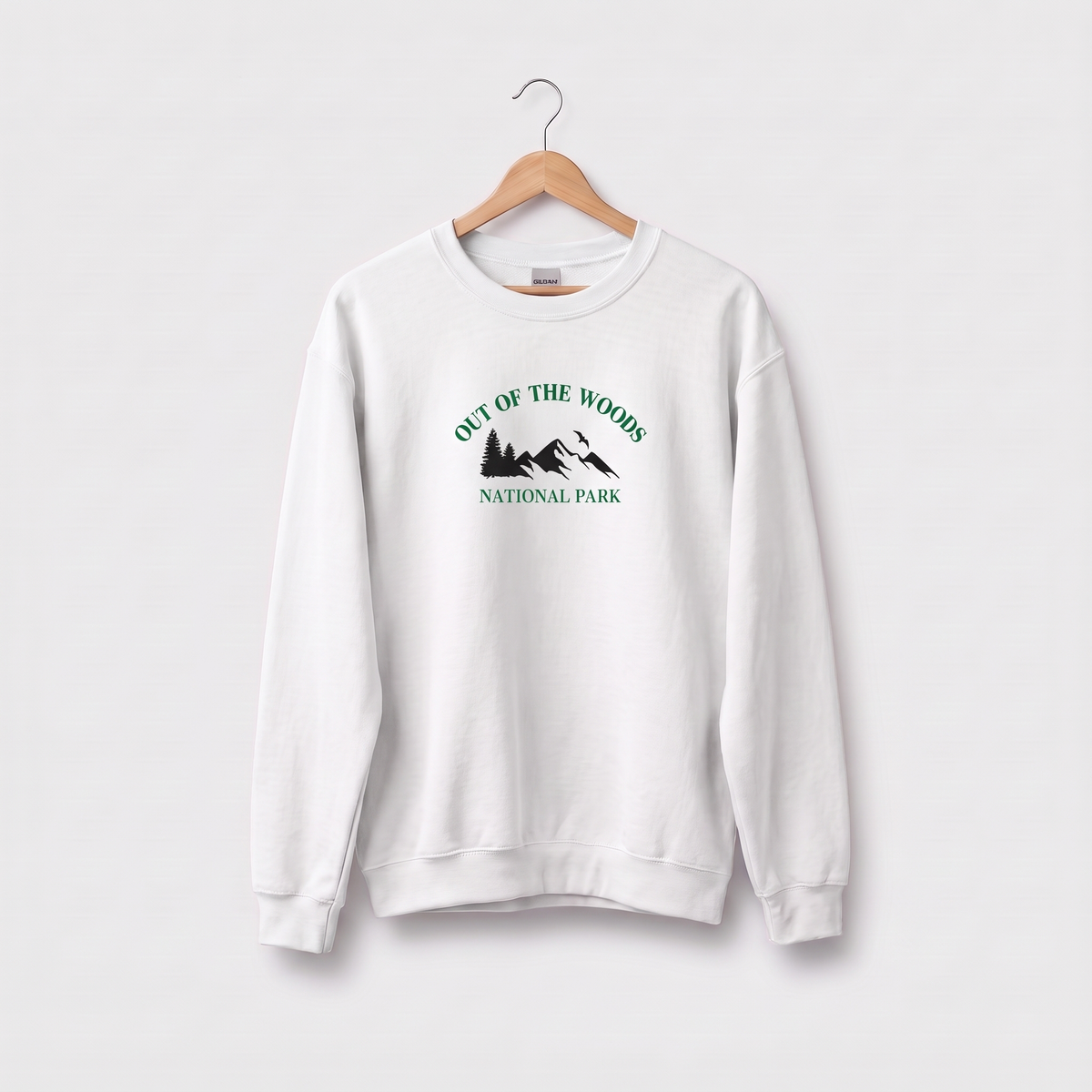 A white crewneck sweatshirt hanging on a wooden hanger against a light gray background. The sweatshirt features a green graphic print on the chest reading 'OUT OF THE WOODS NATIONAL PARK' with a mountain and tree silhouette.