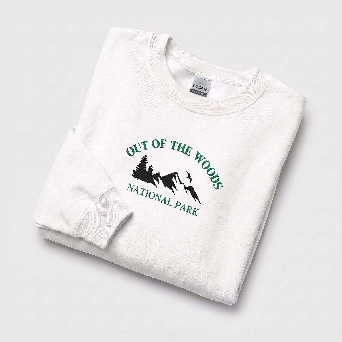 A folded white crewneck sweatshirt on a light gray background, featuring a green graphic print that reads 'OUT OF THE WOODS NATIONAL PARK' with a mountain and tree illustration. The neck label shows the brand Gildan.