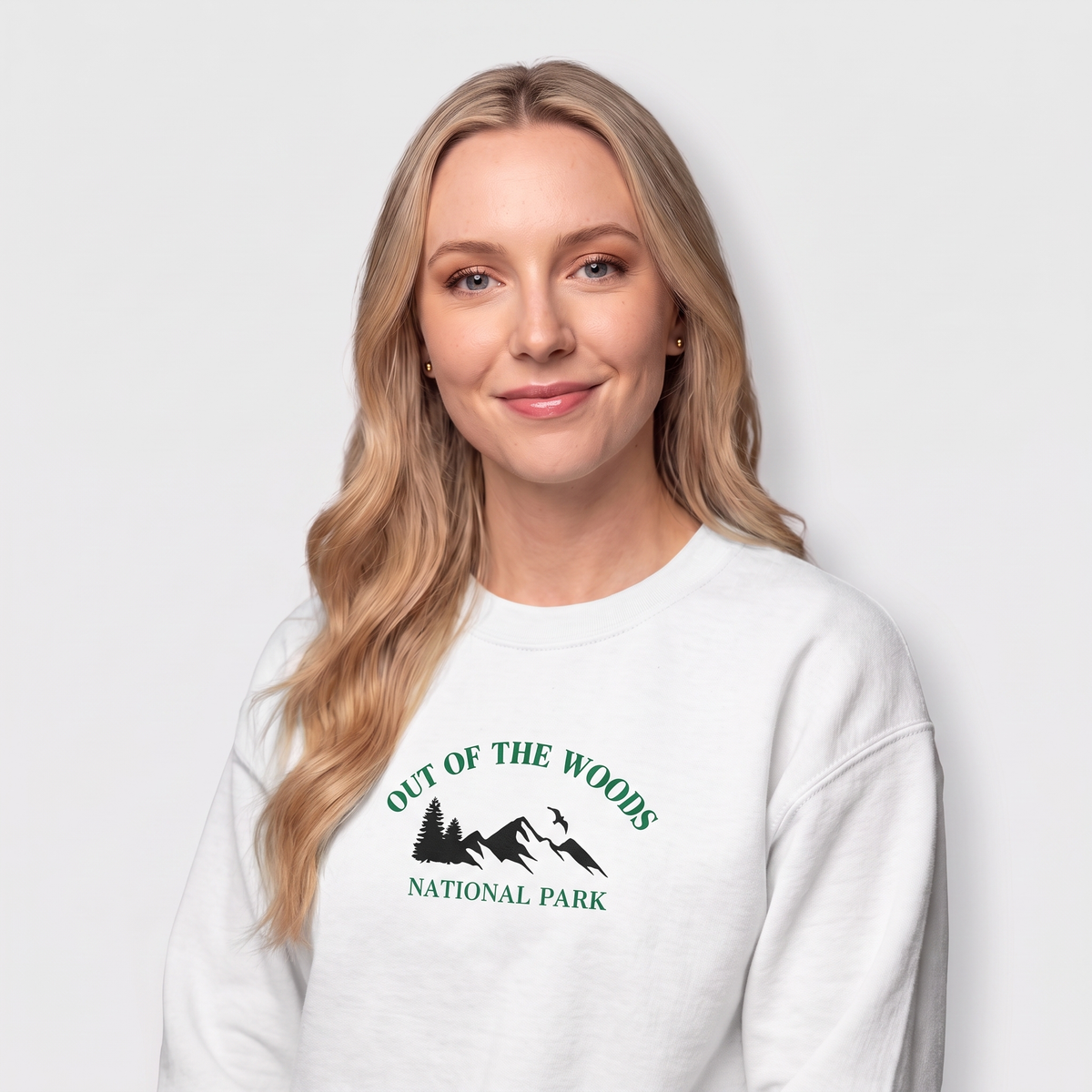 A blonde woman wearing a white crewneck sweatshirt with a green graphic that reads 'OUT OF THE WOODS NATIONAL PARK' with a mountain and tree design, set against a light gray studio background.