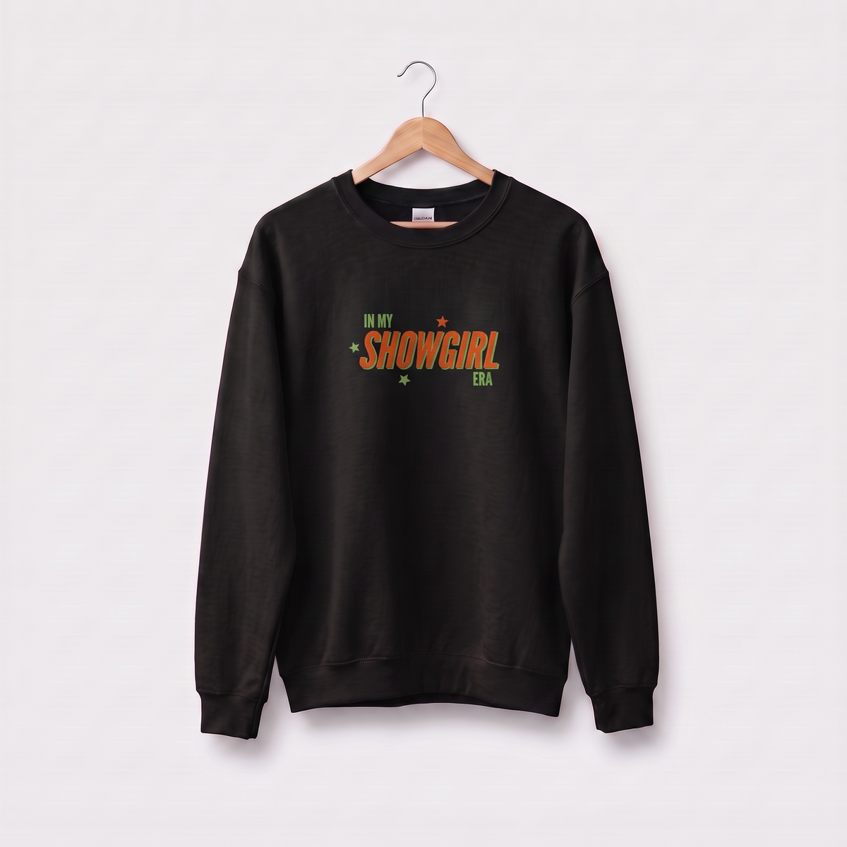 A black crewneck sweatshirt hanging on a wooden hanger against a light gray background. The sweatshirt features a graphic on the chest that reads 'IN MY SHOWGIRL ERA' in orange and green lettering.