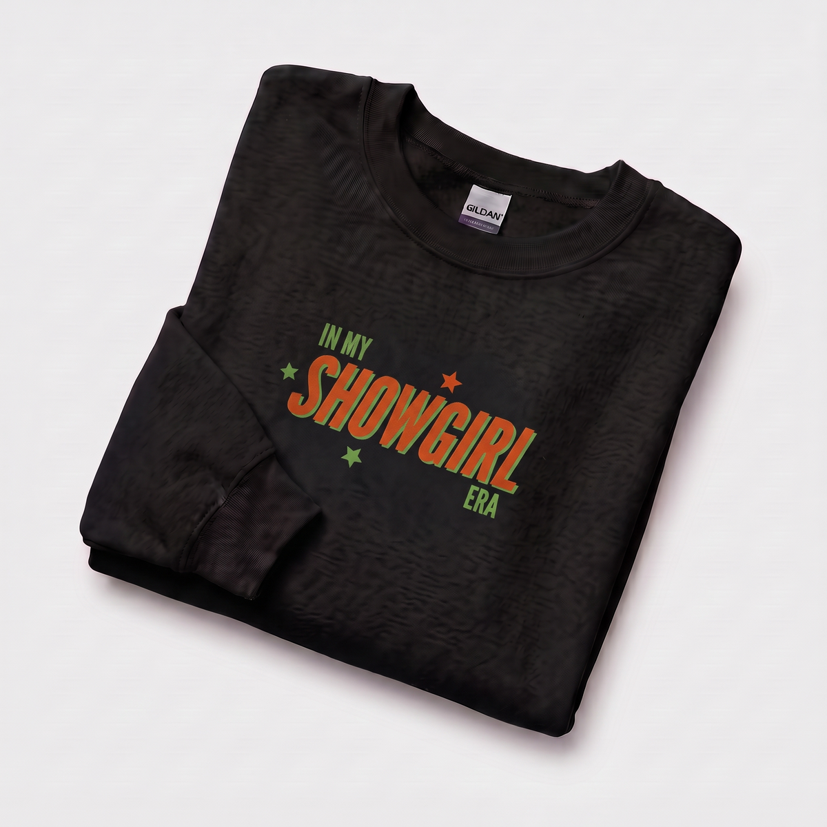 A folded black crewneck sweatshirt with a textured pattern, featuring a colorful graphic print on the front that reads 'IN MY SHOWGIRL ERA' in orange and green with small star accents. The neck label shows the brand Gildan.