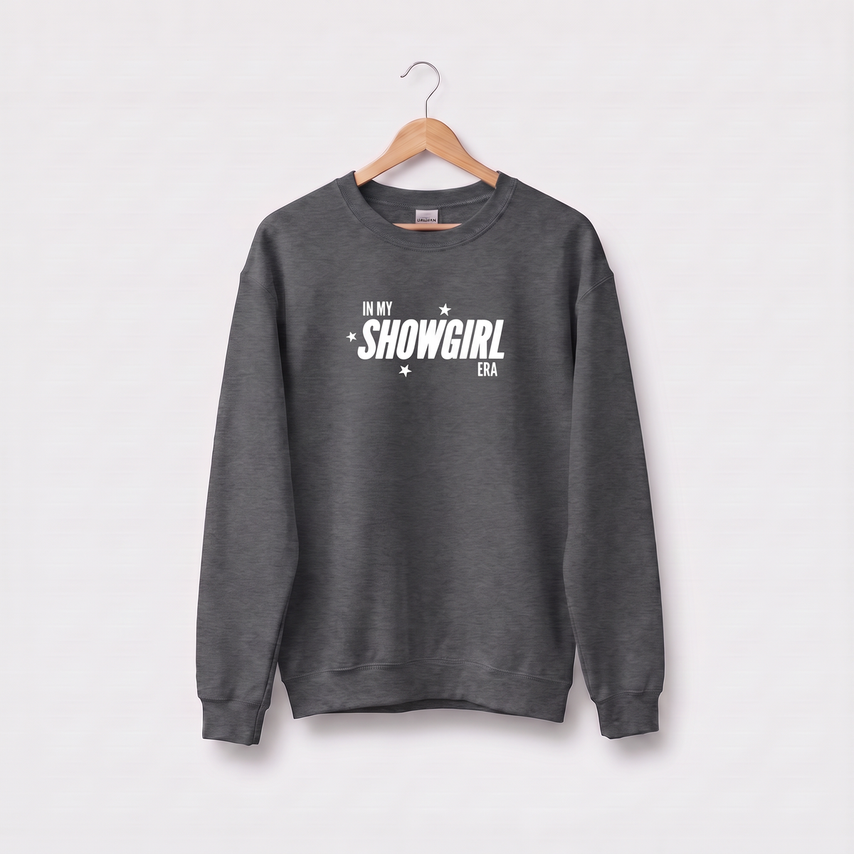 A dark gray crewneck sweatshirt hanging on a wooden hanger against a light gray background. The sweatshirt features the text 'IN MY SHOWGIRL ERA' printed in white on the chest, accented with small white stars.