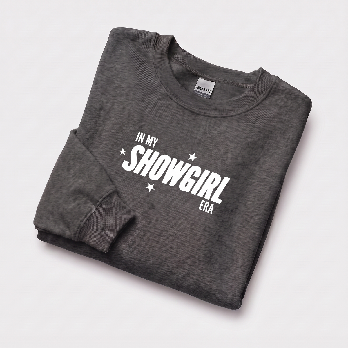 A folded dark gray heather crewneck sweatshirt with 'IN MY SHOWGIRL ERA' printed in white across the chest, displayed on a light gray background.