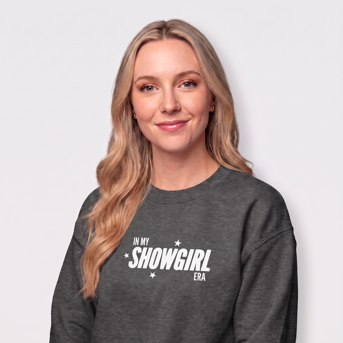 A woman with blonde hair wearing a dark gray heather crewneck sweatshirt with the white text 'IN MY SHOWGIRL ERA' printed on the chest, set against a plain light gray background.