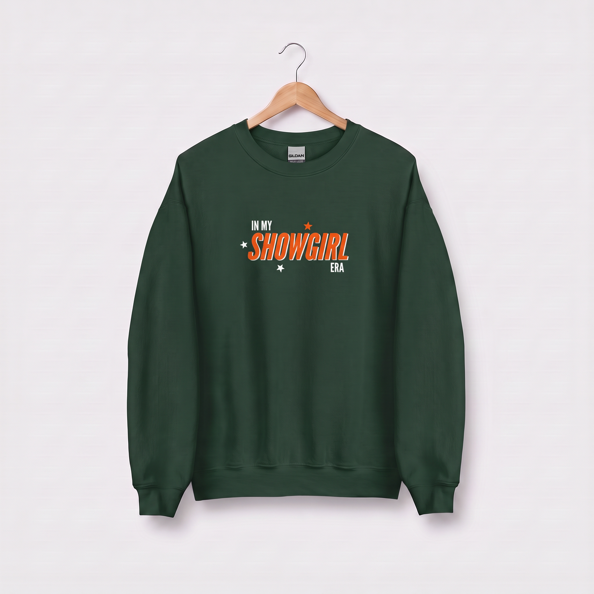 A dark green crewneck sweatshirt hanging on a wooden hanger against a light gray background. The sweatshirt features a graphic on the chest that reads 'IN MY SHOWGIRL ERA' in orange and white lettering with small star and heart accents.