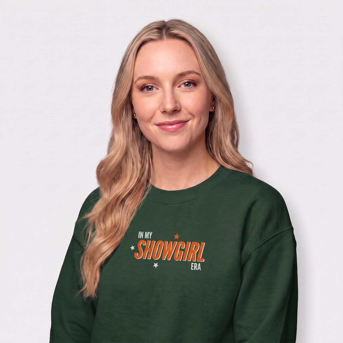 A woman with long blonde hair wearing a dark green crewneck sweatshirt with the text 'IN MY SHOWGIRL ERA' printed in orange and white on the chest, set against a plain light gray background.