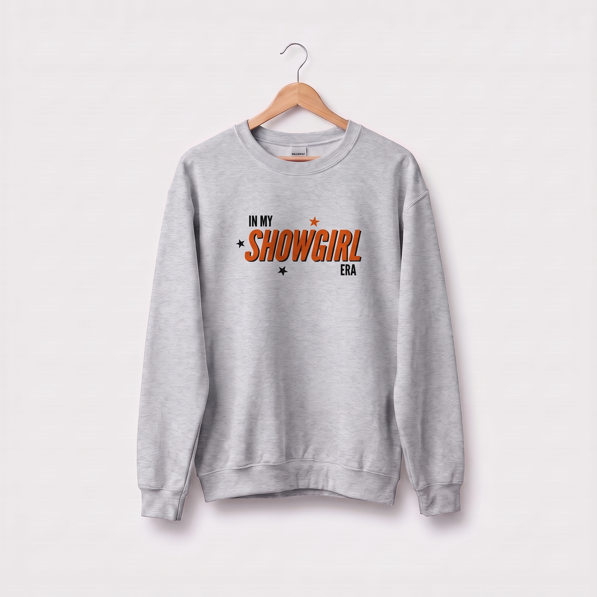 A light gray crewneck sweatshirt hanging on a wooden hanger against a light gray background. The sweatshirt features a graphic on the chest that reads 'IN MY SHOWGIRL ERA' in orange and black text with small star accents.