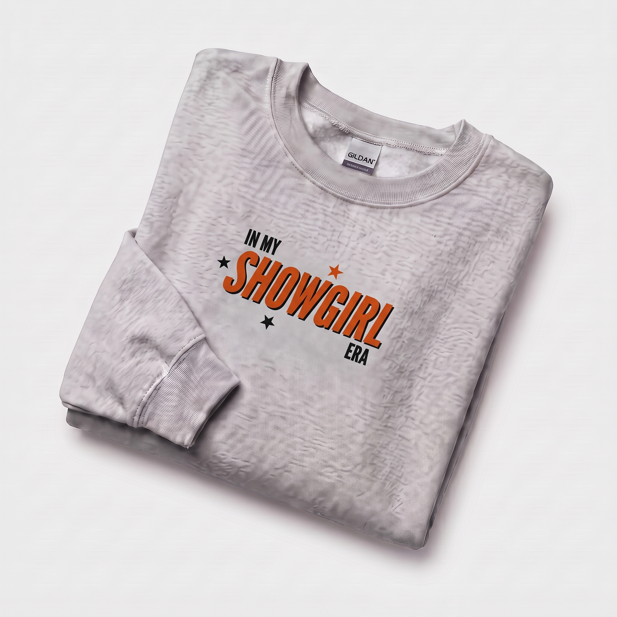 A folded light gray sweatshirt with a textured finish, featuring the graphic text 'IN MY SHOWGIRL ERA' in orange and black on the chest, displayed on a clean white background.