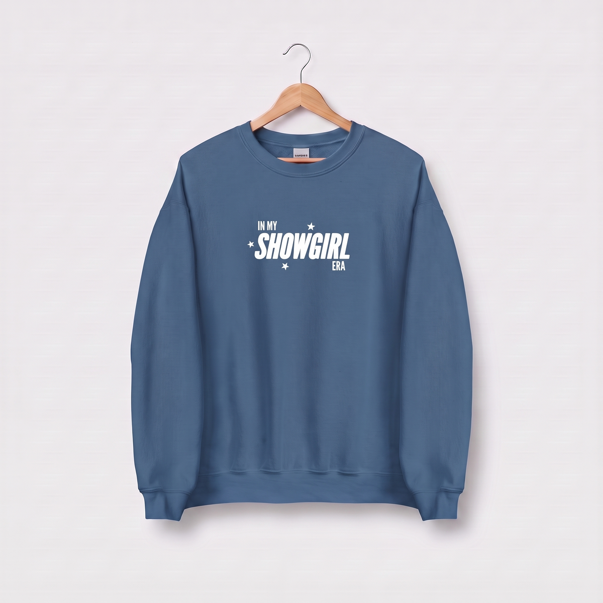 A dusty blue crewneck sweatshirt hanging on a wooden hanger against a light gray background. The sweatshirt features the text 'IN MY SHOWGIRL ERA' printed in white across the chest.