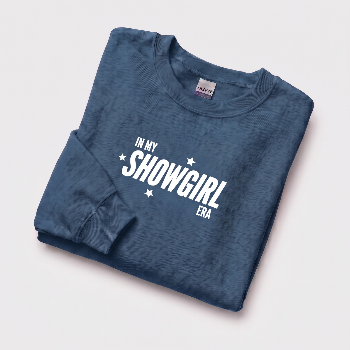 A folded slate blue long-sleeve t-shirt with a textured pattern, featuring the white graphic text 'IN MY SHOWGIRL ERA' with small white stars, displayed on a light gray background.