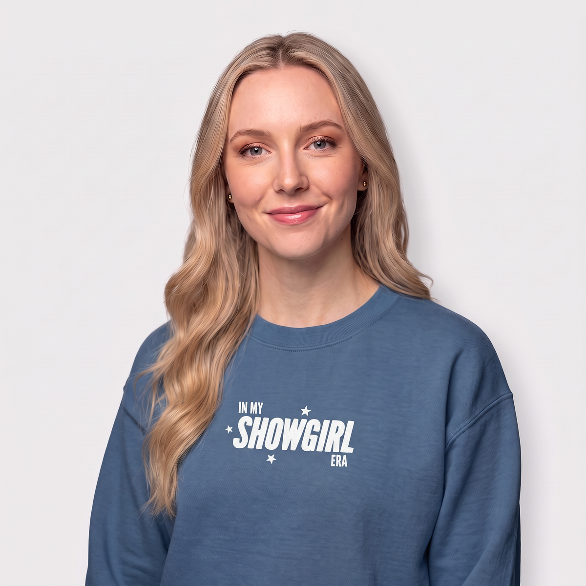 A woman with long blonde hair wearing a slate blue crewneck sweatshirt with white text that reads 'IN MY SHOWGIRL ERA' against a light gray background.